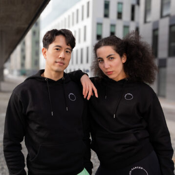 To personer i svarte hettegensere med minimalistisk logo poserer selvsikkert i et urbant miljø. Den ene har lysegrønne shorts, den andre svarte leggings med "FUCK CANCER" trykt på låret.